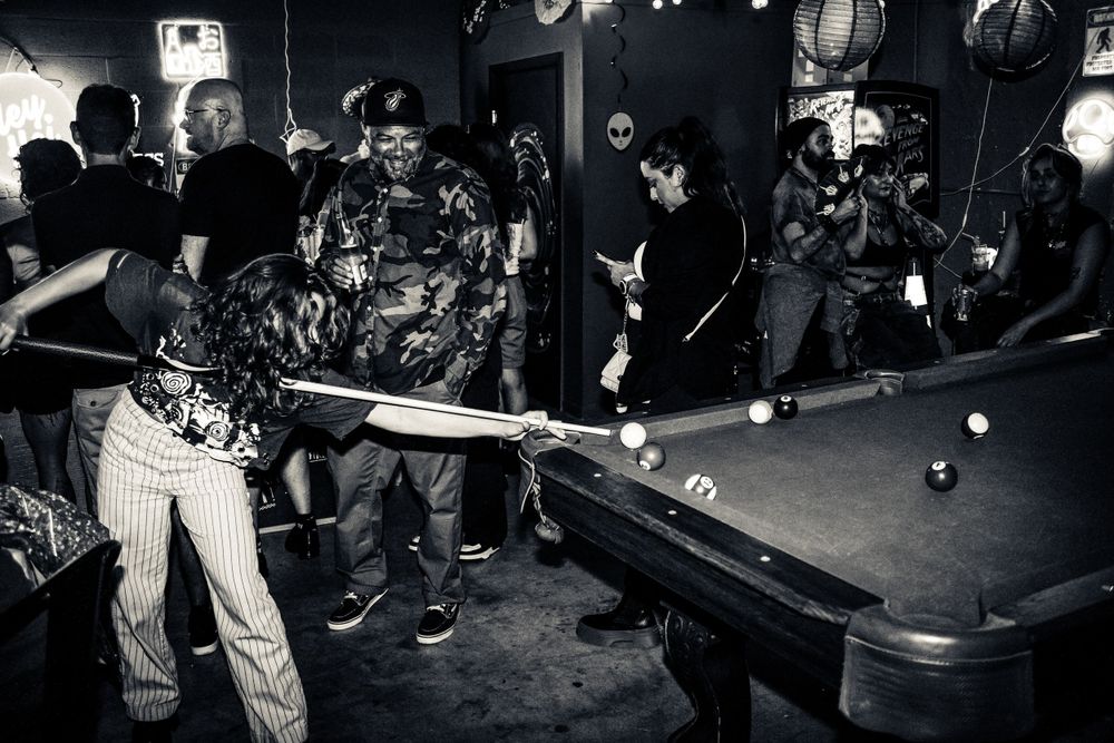 Black and white photograph of people playing pool in a dimly lit bar setting.