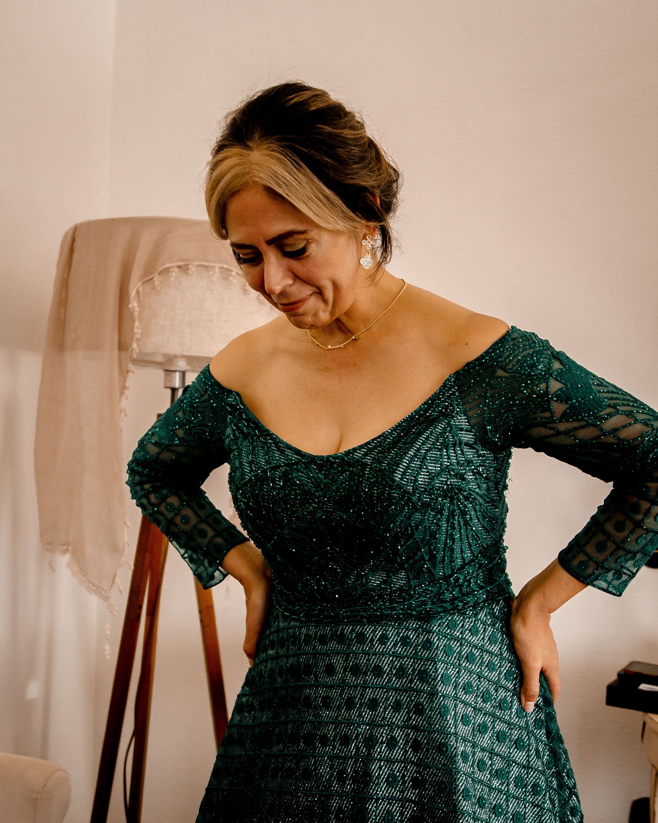 A person wearing an elegant dark green lace evening gown with long sleeves stands in a room with soft lighting.