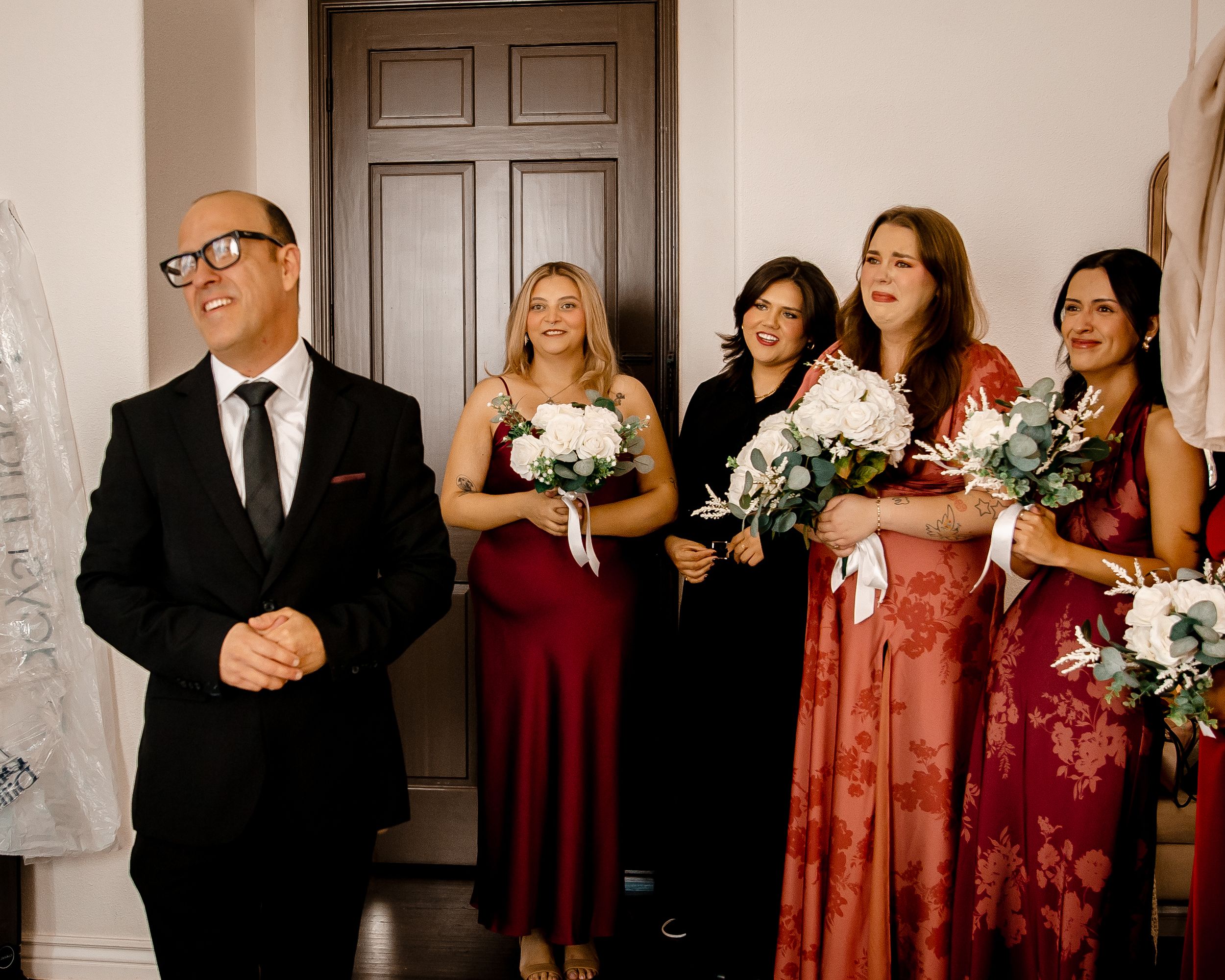 A wedding party in formal attire stands together holding white bouquets against a neutral wall with a dark wooden door.