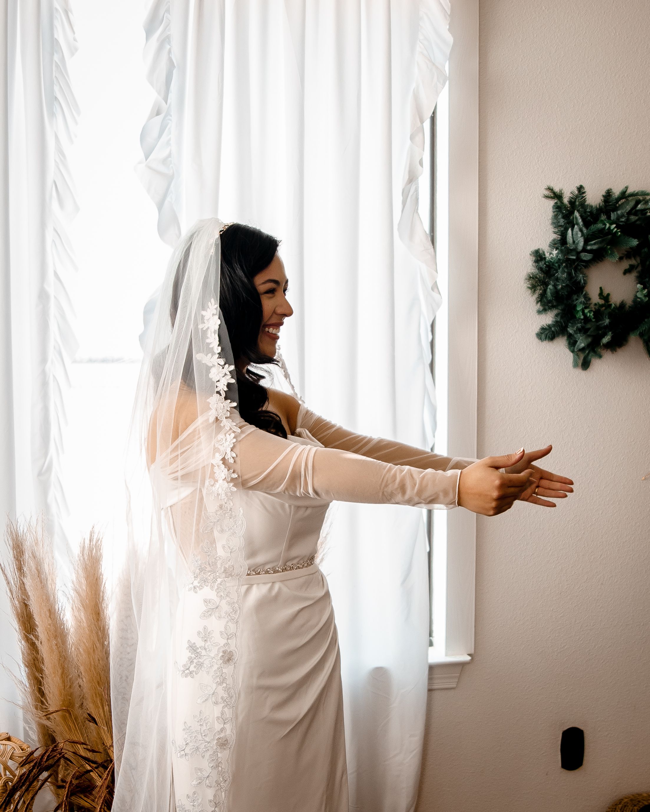 A person in a white dress reaches toward sunlit curtains with pampas grass decor creating a dreamy bridal atmosphere.