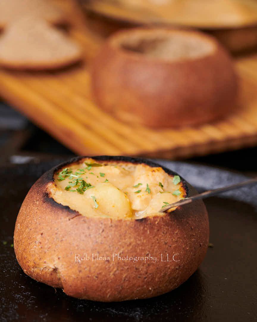 Bread bowls being filled with soup.  Bread bowls in background are on cutting board.