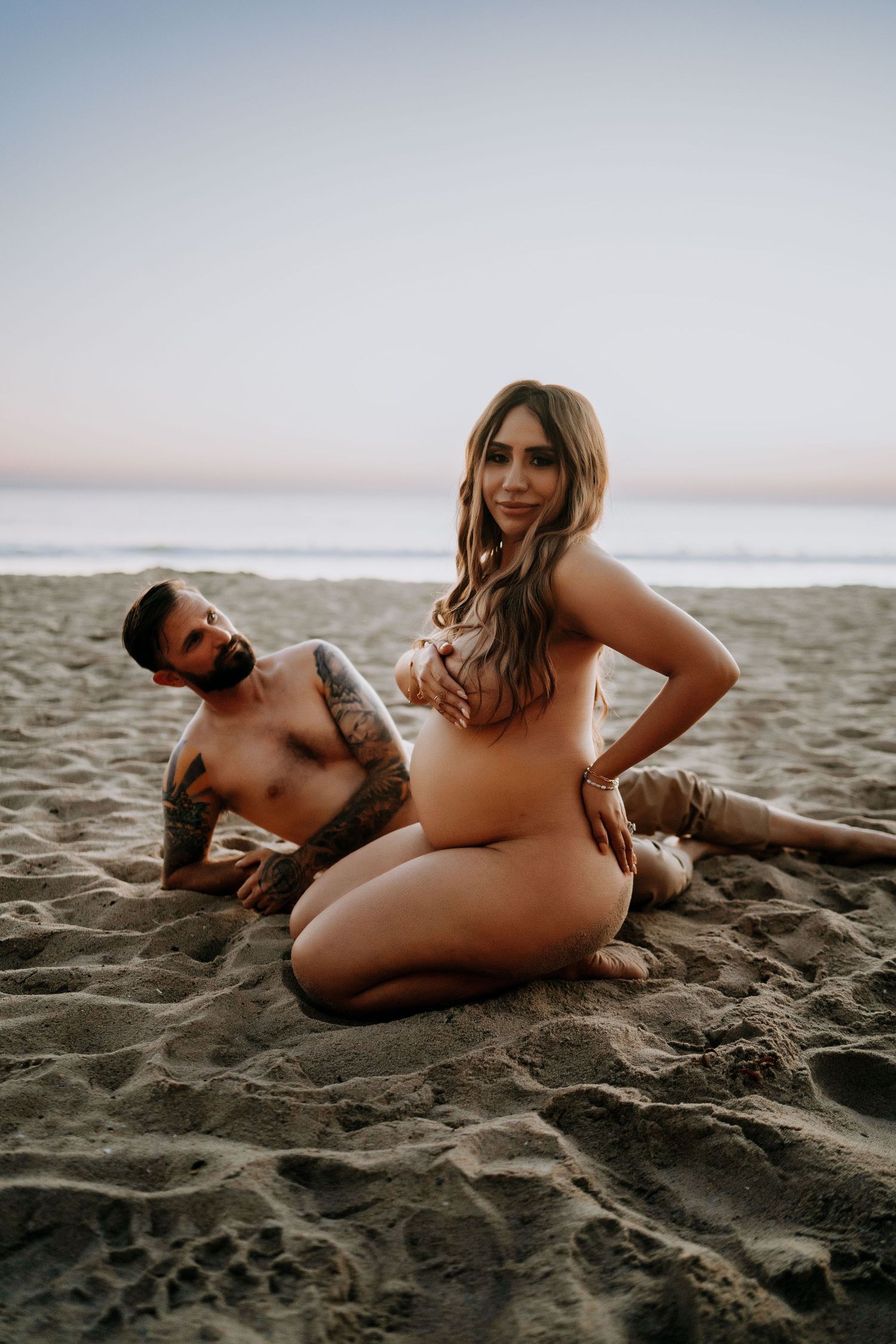 Nude pregnant woman and tattooed shirtless man pose together on sandy beach at sunset.