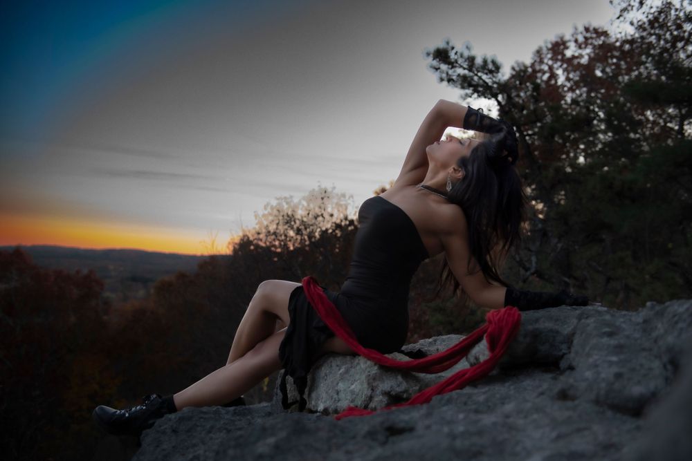 Artistic photo of a person in black dress posing on a rocky cliff edge at sunset with dramatic sky backdrop.