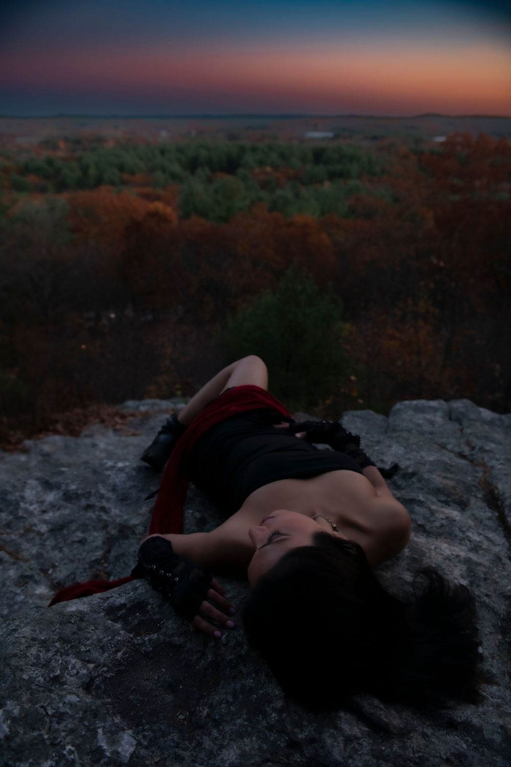 Figure in black dress lying on rocky outcrop against autumn forest vista at dusk.