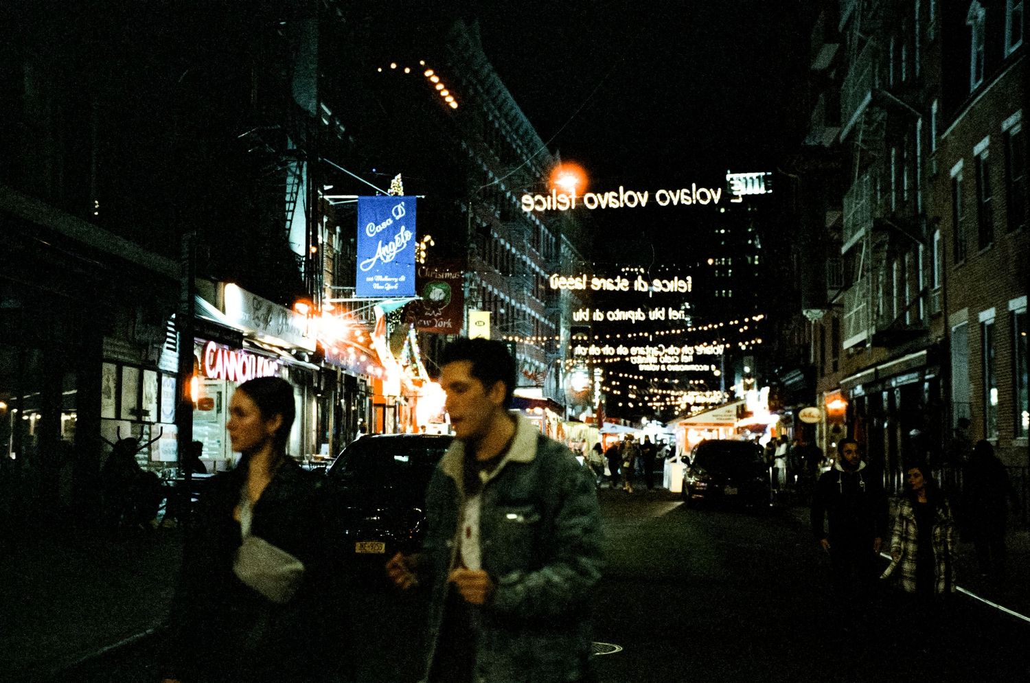 City street at night with illuminated storefront signs and pedestrians.