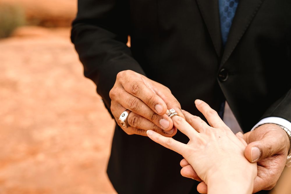 Close-up of hands exchanging wedding rings against formal attire.