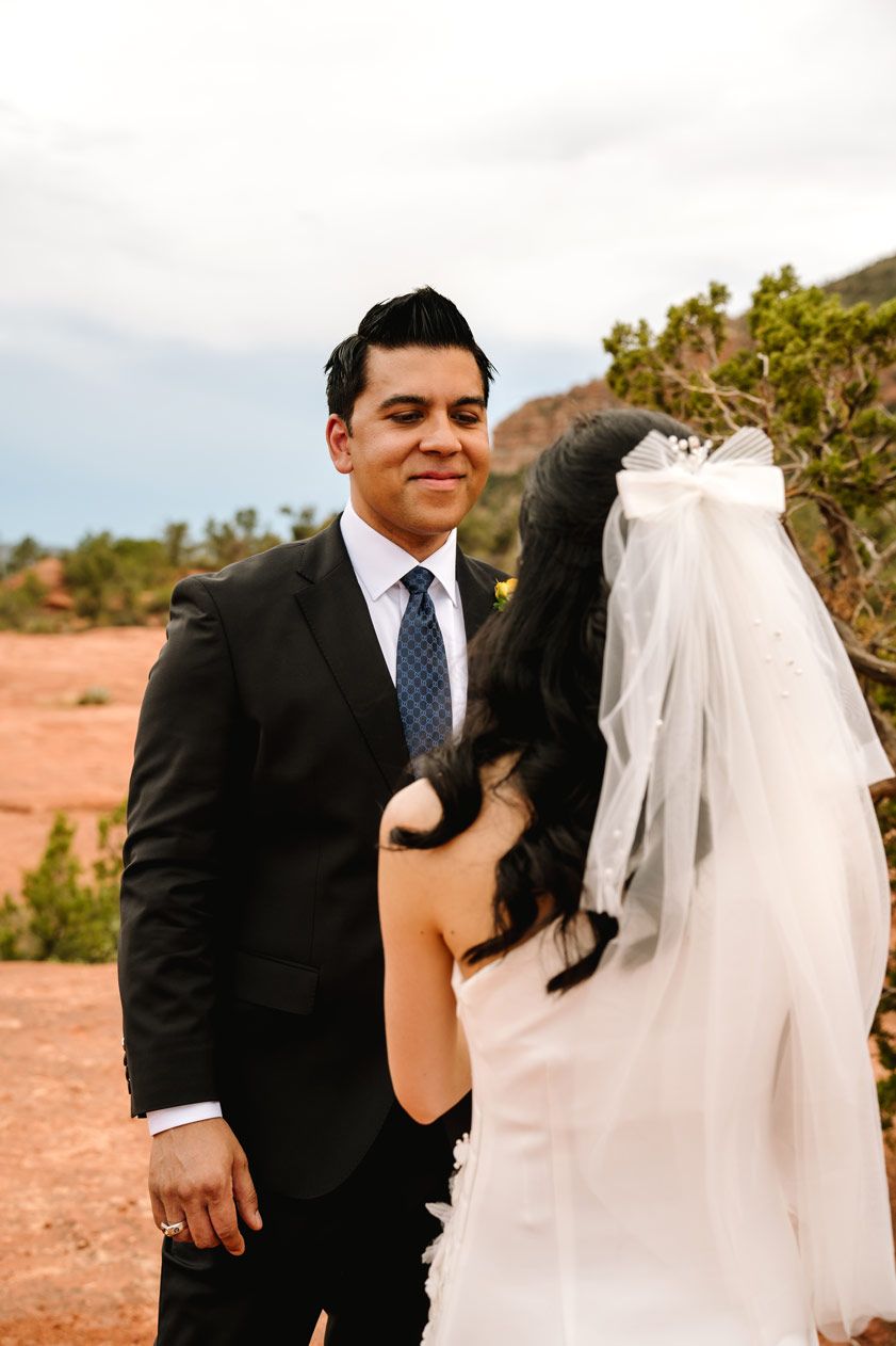 A couple in formal wedding attire stands against a Sedona desert landscape with rocky cliffs.