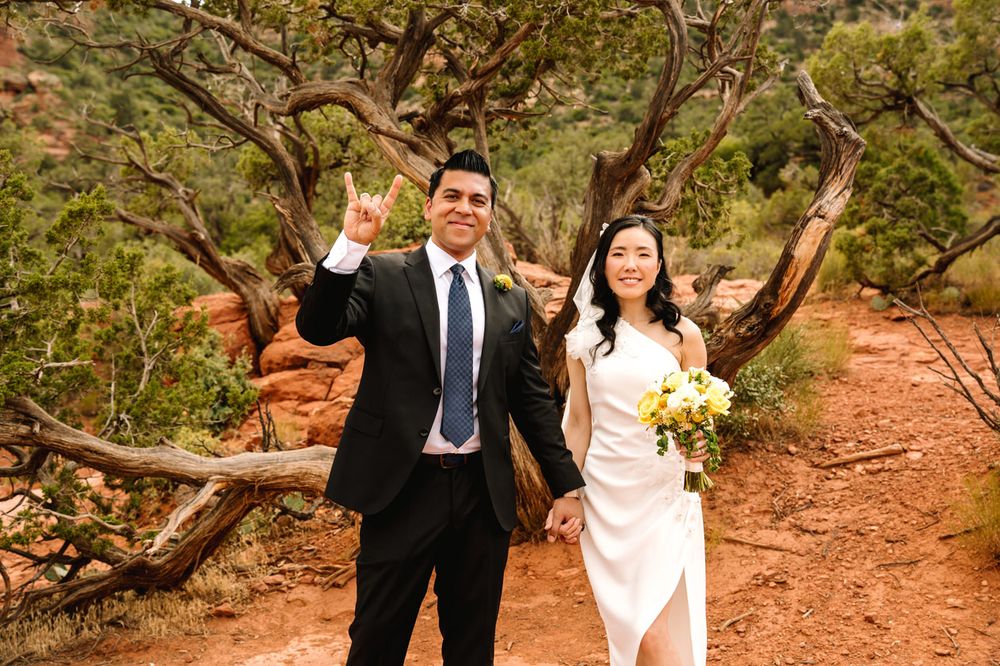 Newlyweds celebrate with raised arms on a desert trail surrounded by Sedona red rocks and desert vegetation.