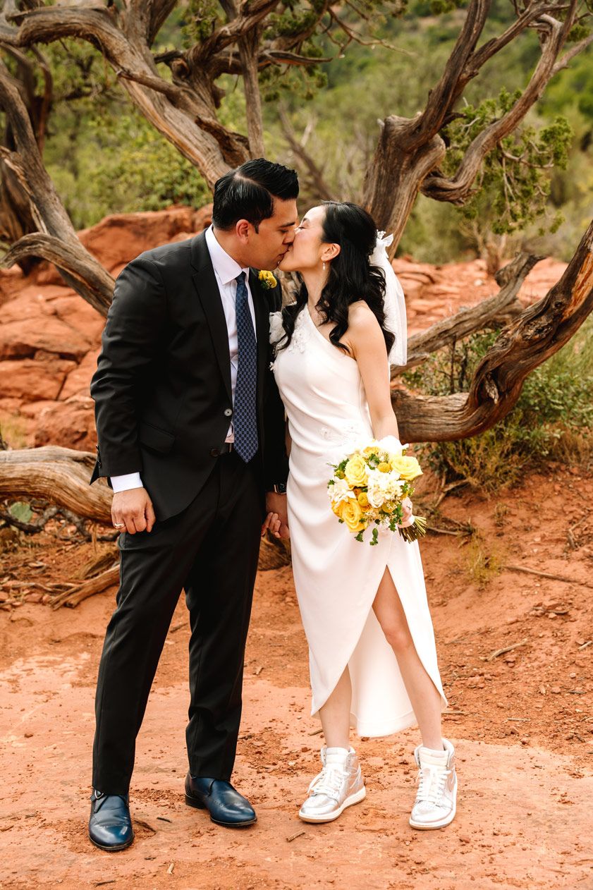 A couple shares a kiss in wedding attire with the bride wearing sneakers against Sedona desert scenery.
