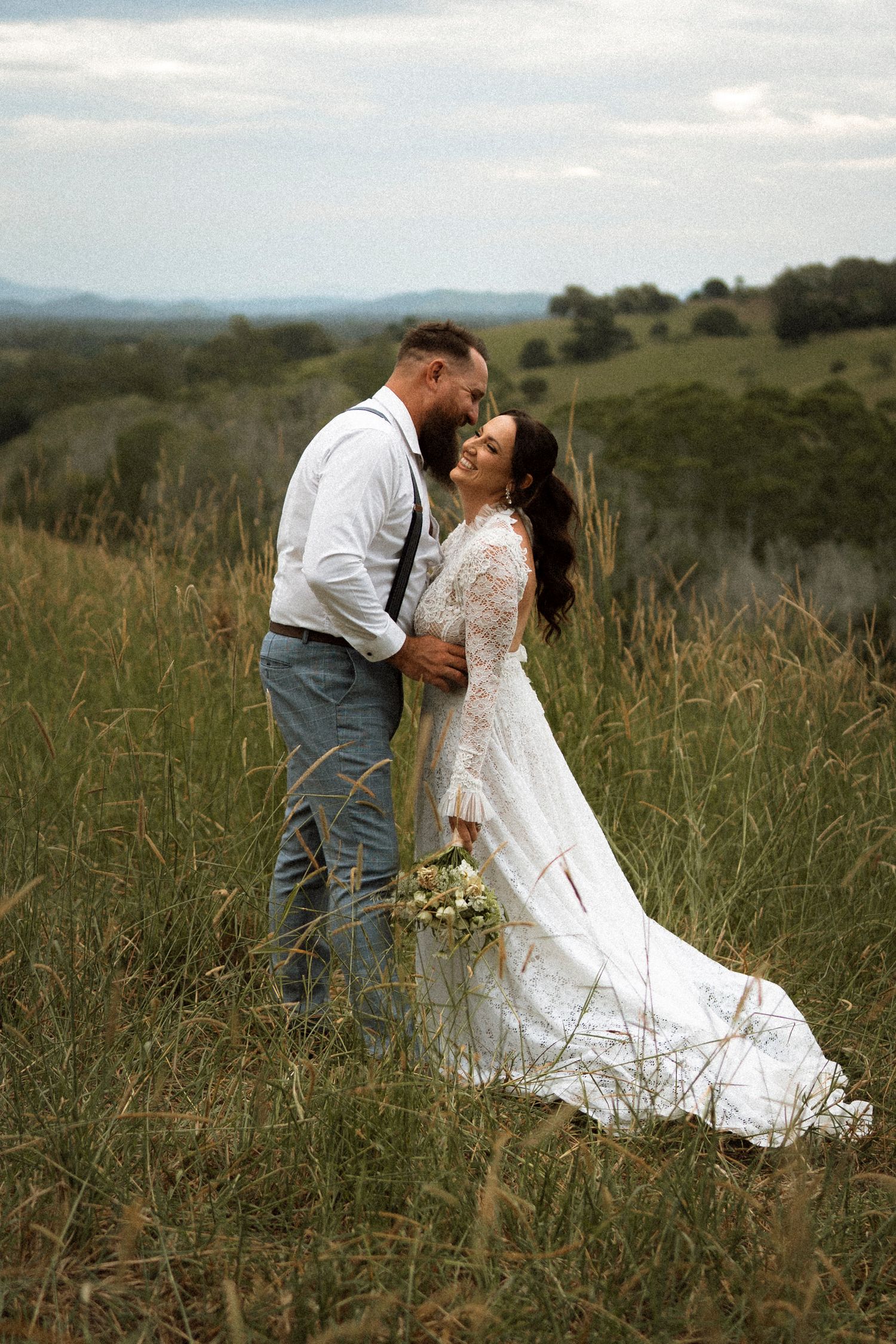 A couple in wedding attire embraces in a grassy field, bride in lace gown holding bouquet, scenic hills in background.