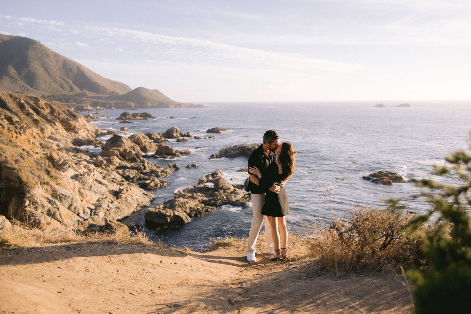 Two people embrace while standing on a cliff overlooking a rocky Pacific coastline during golden hour.