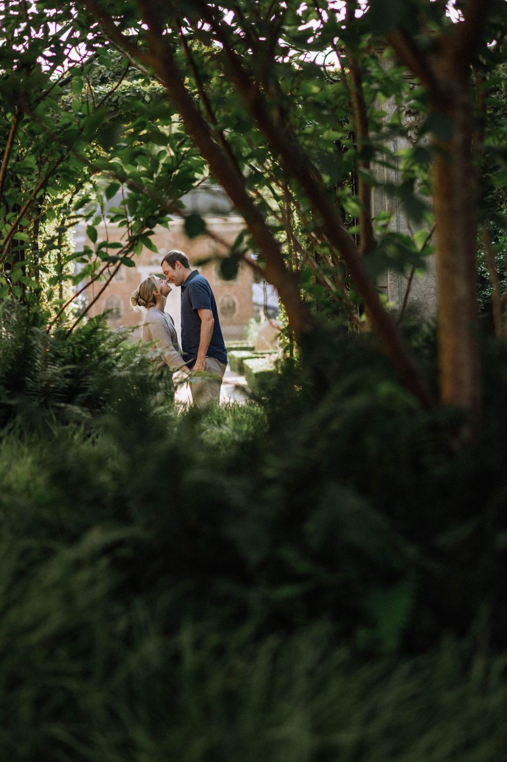 Couple embracing in a lush garden setting with dappled sunlight filtering through tree branches.