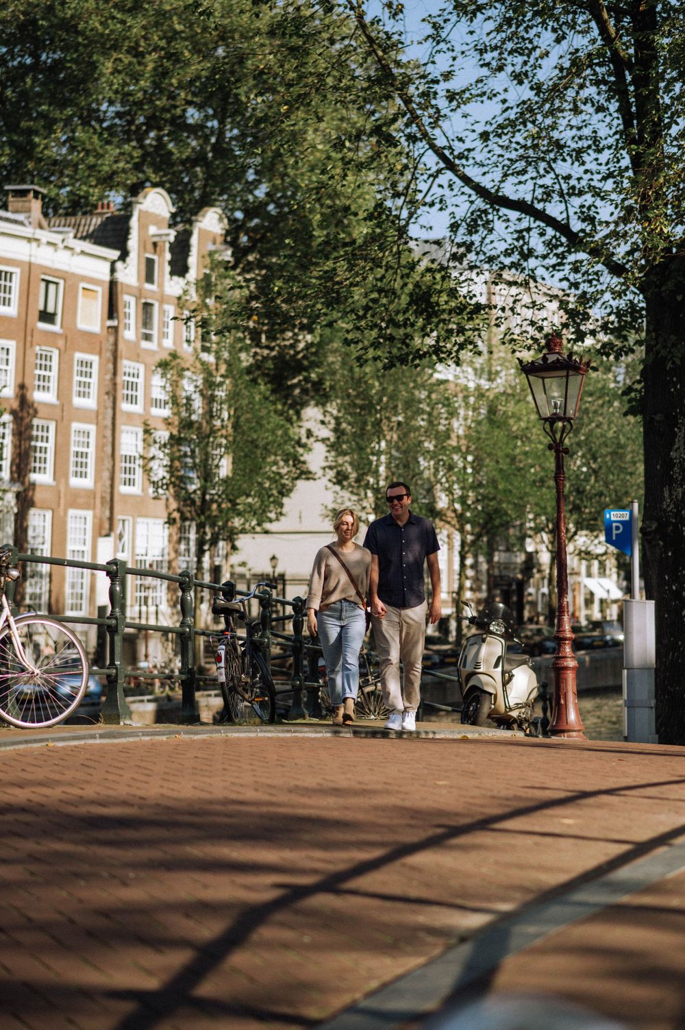 A sunny day in Amsterdam with people walking past traditional brick buildings and trees.