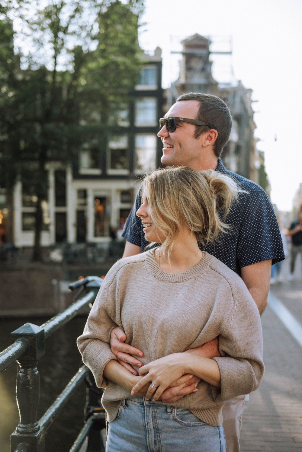 Couple embracing on Amsterdam bridge with classic Dutch architecture in background.