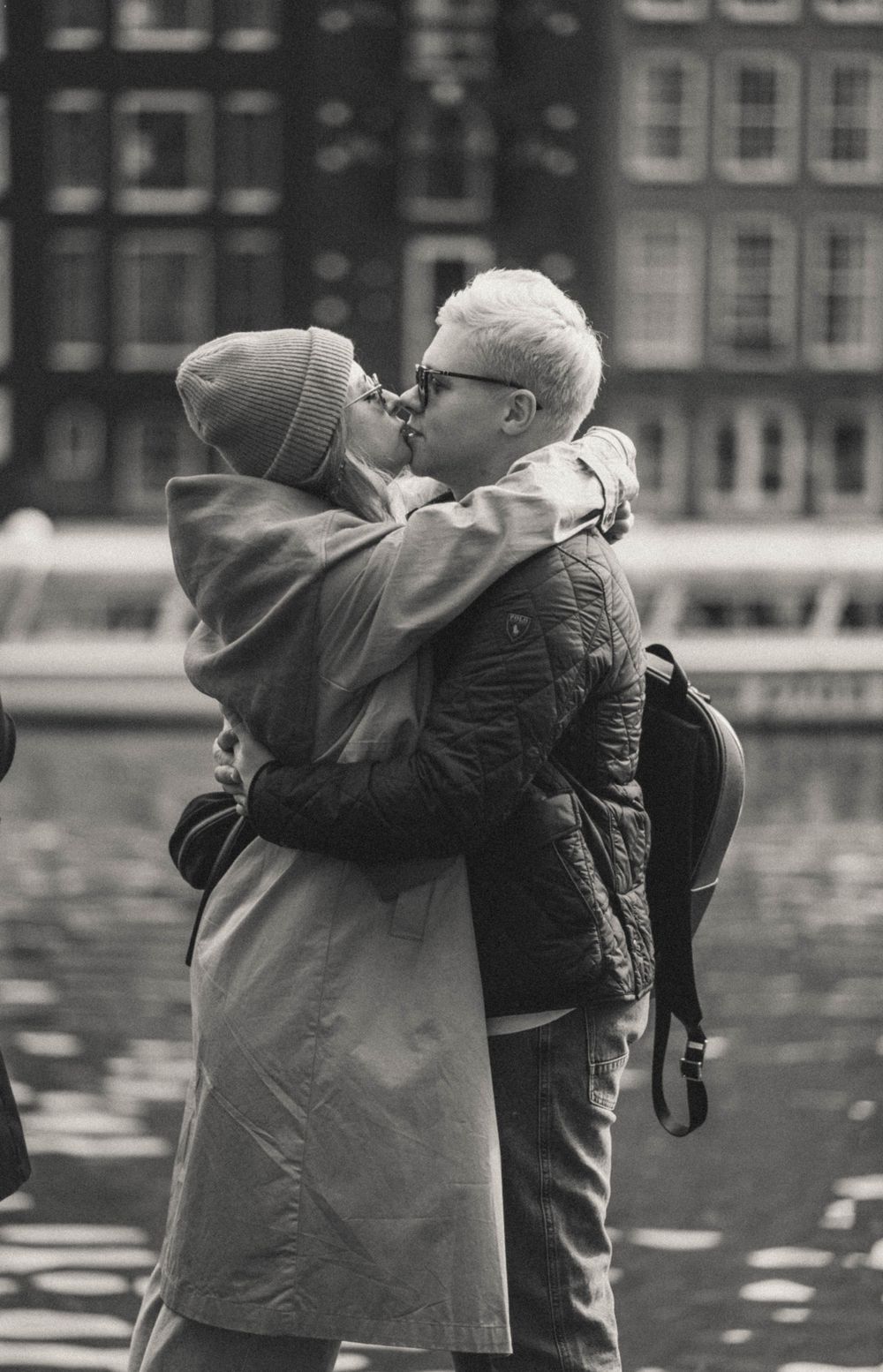 Black and white photo of couple embracing by Amsterdam canal with reflections on water.
