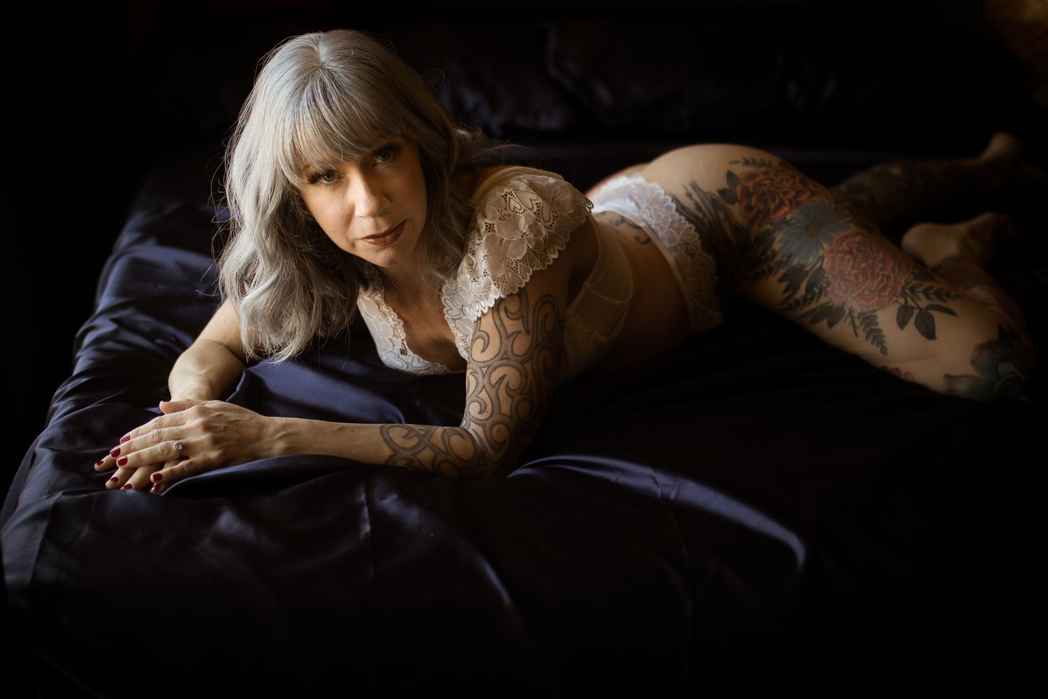 A mature woman celebrates with a birthday boudoir shoot in Fort Worth wearing white lingerie while lounging on a dark luxurious bed.