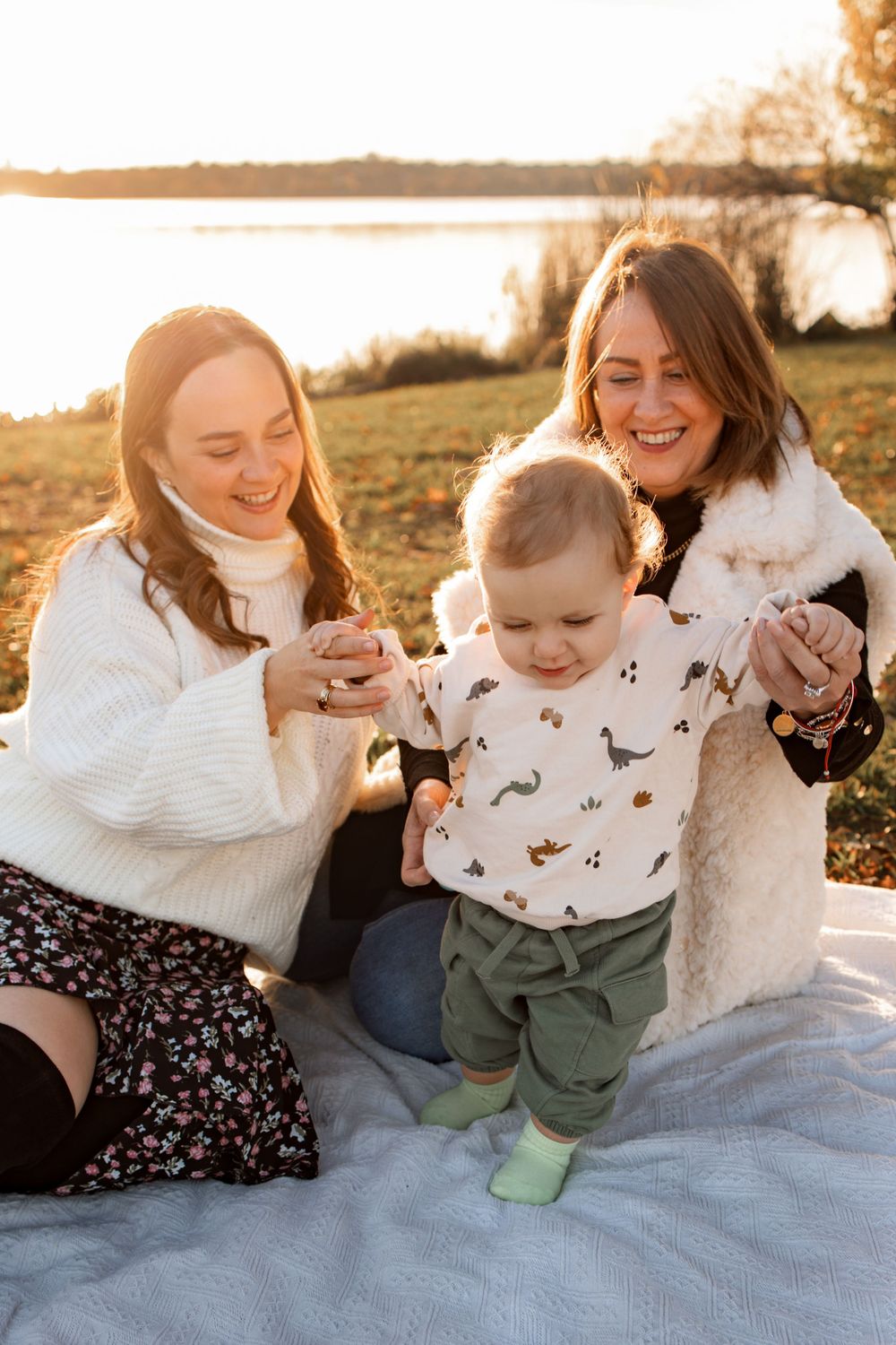 Family enjoying playful moment with toddler on white blanket by lake at sunset.