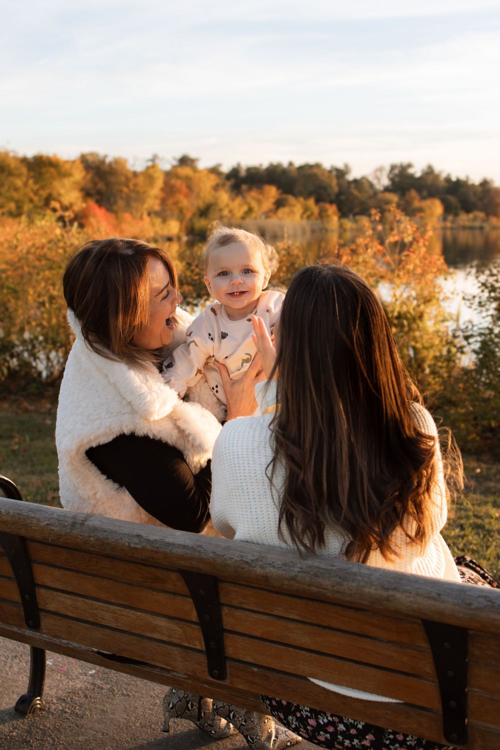 Three people sit together on a park bench at sunset with autumn foliage and a lake in the background.