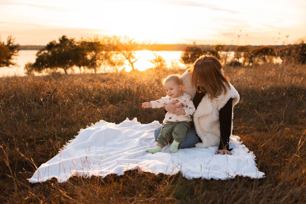 Two people and baby sitting on white blanket during golden sunset in grassy field.
