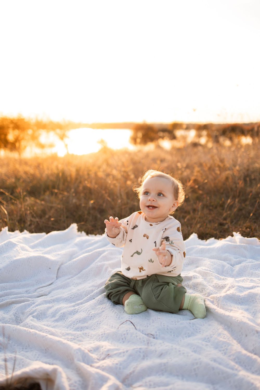 Baby sitting on white blanket outdoors during sunset wearing printed shirt and green pants.