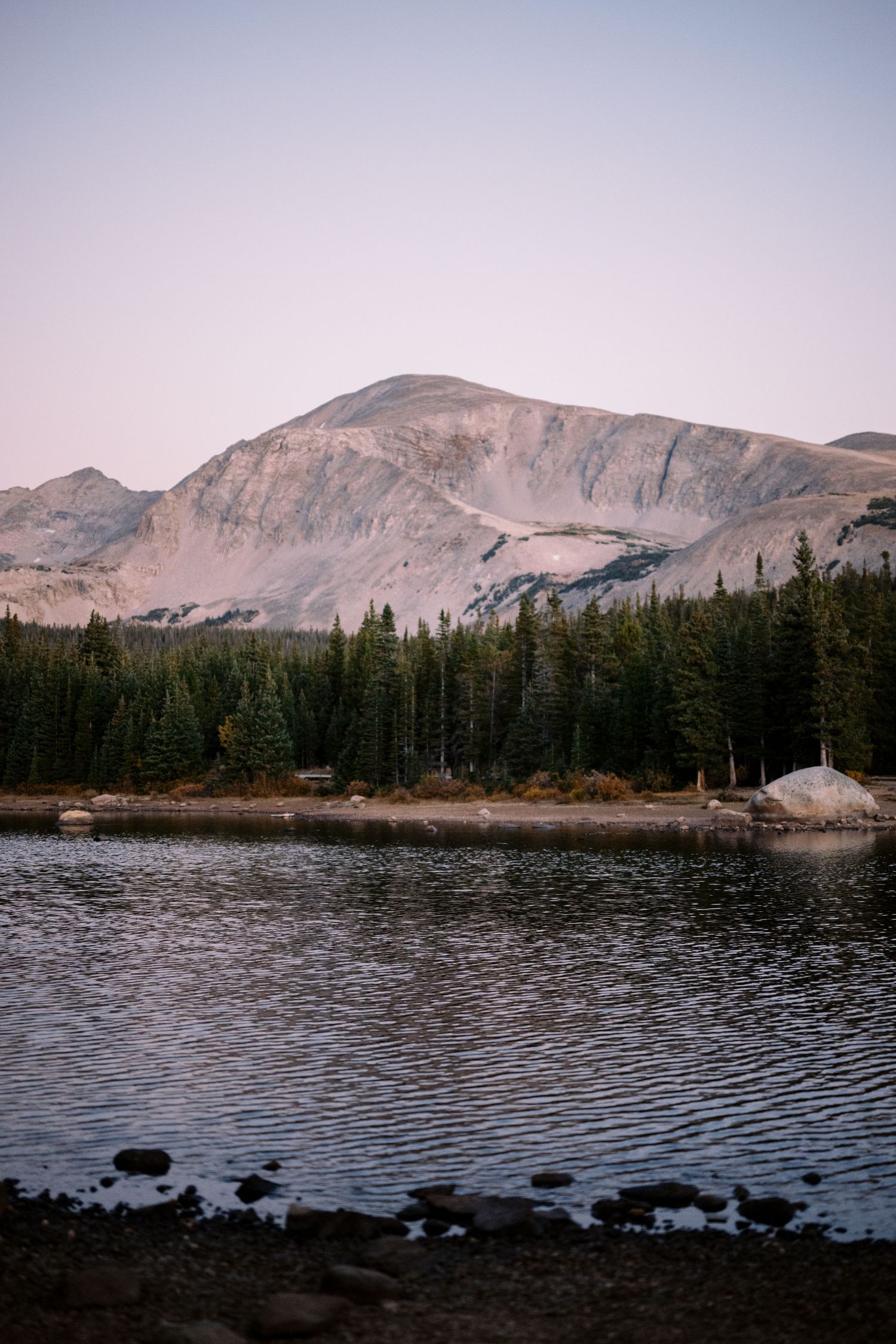 Scenic mountain lake surrounded by evergreen forest with rocky peaks and purple dusk sky in the background.