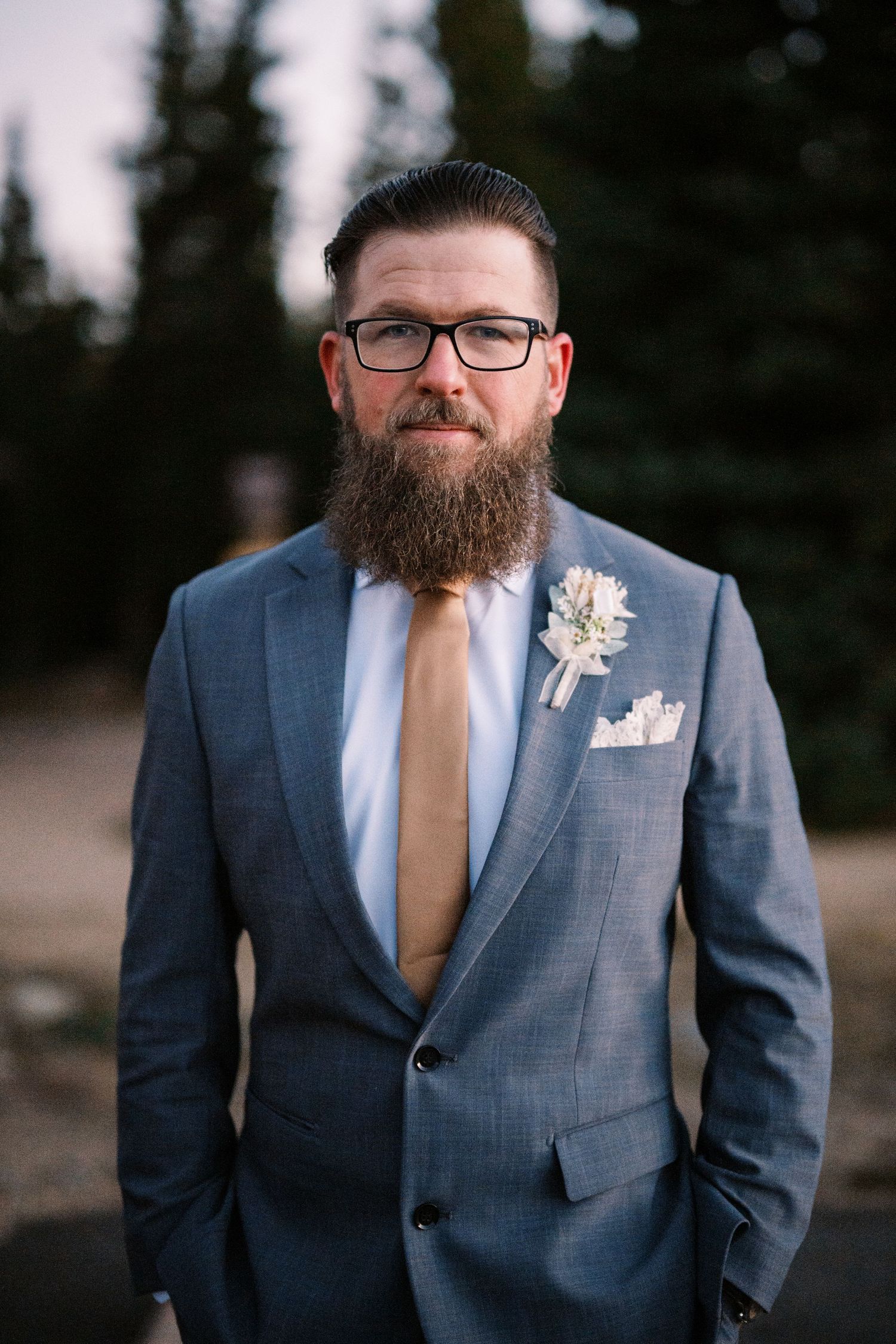 A groom in a blue suit with a brown tie and full beard wears glasses and a boutonniere at an outdoor wedding.