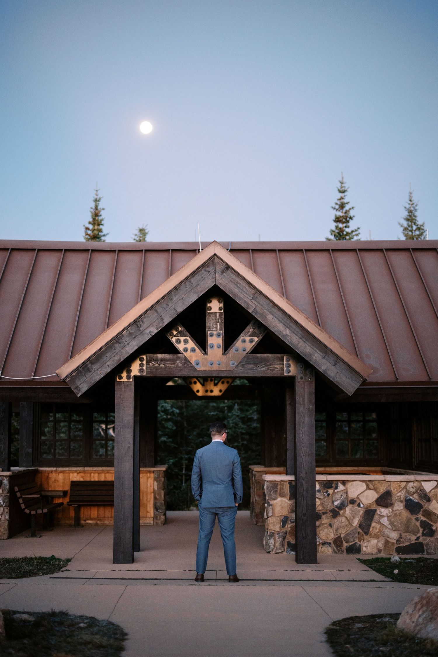 A figure in a blue suit stands at the entrance of a rustic mountain lodge with stone walls and timber beams at dusk.
