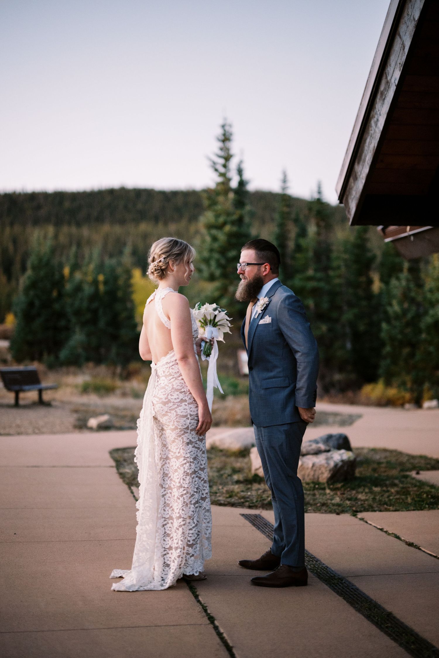 A couple stands together at a mountain venue, with the bride in a lace gown and groom in navy suit against evergreen scenery.
