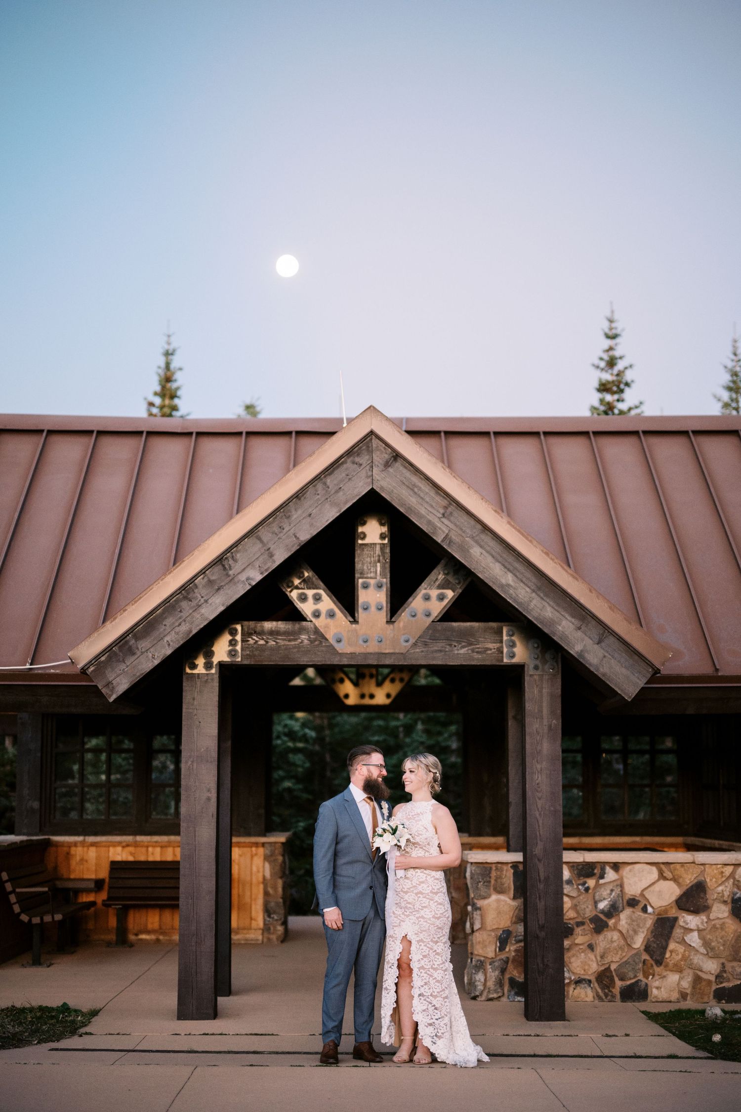 Couple stands in front of rustic timber entrance of mountain lodge at dusk with full moon in blue sky.