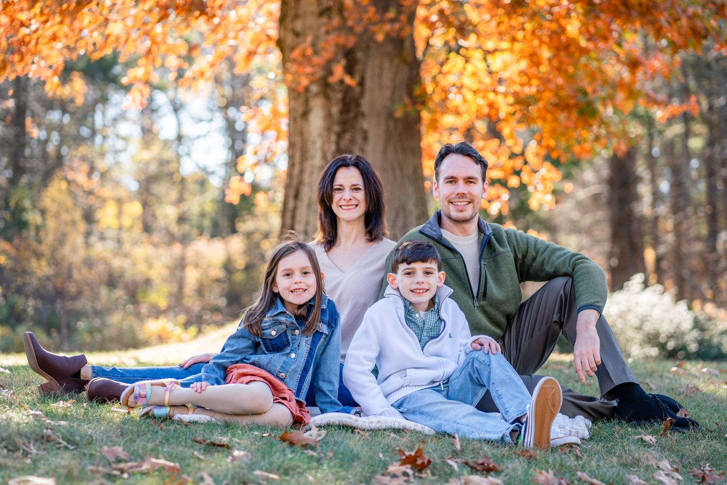 Family sitting together on grass during autumn with colorful fall foliage in background.