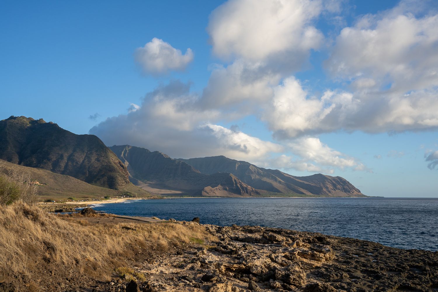 Rocky coastline with dramatic mountains and blue waters under puffy clouds.
