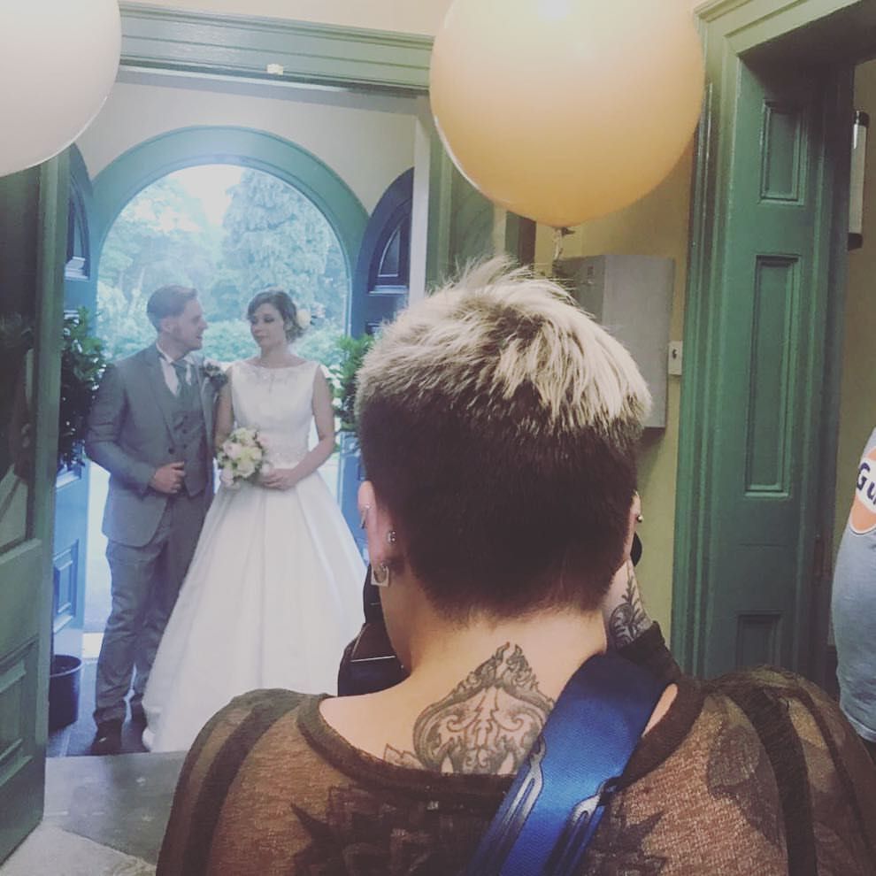 Two people stand in a wedding ceremony setting under an archway, viewed through the shoulder of a guest with a balloon.