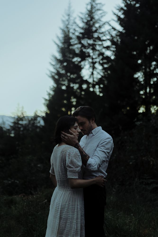 A couple embraces in a forest clearing at dusk surrounded by evergreen trees.