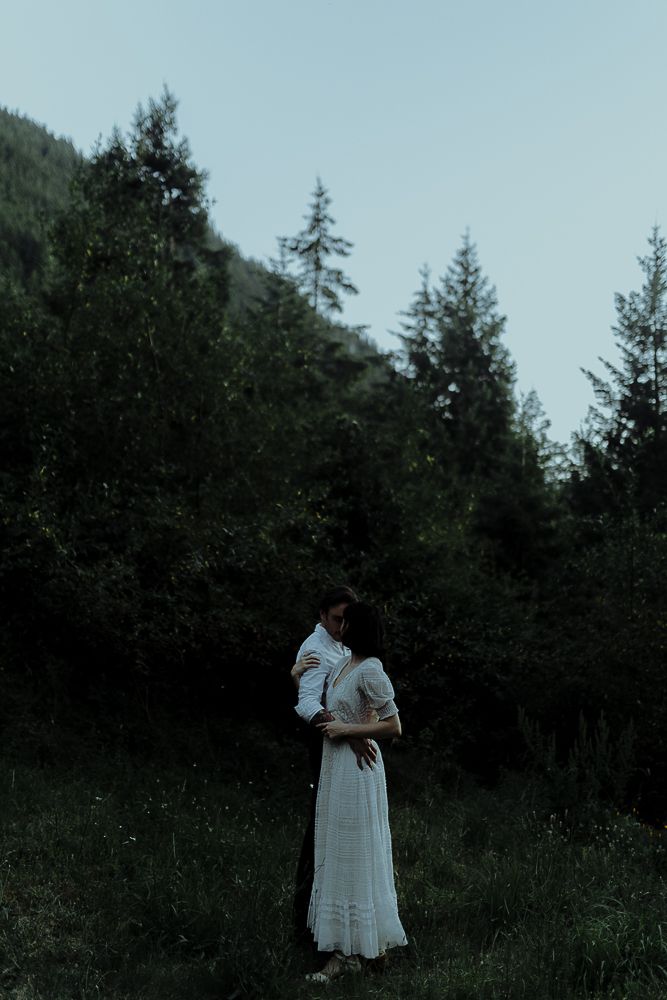 Two figures in white stand together against an evergreen forest backdrop.