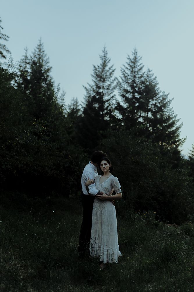 A couple stands together in a meadow with evergreen trees in the background at twilight.