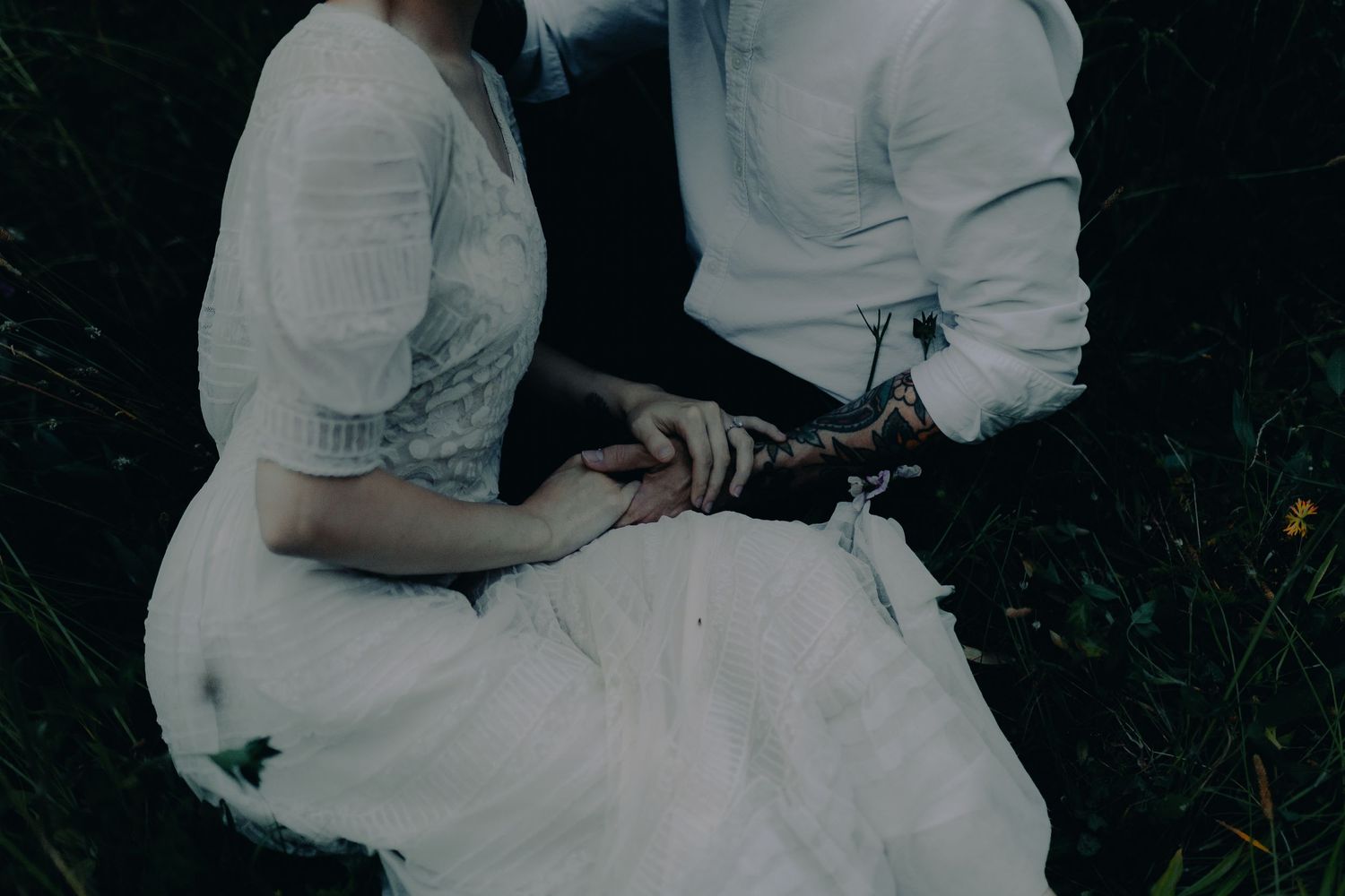 A couple in white clothing embraces intimately in a moody outdoor setting.