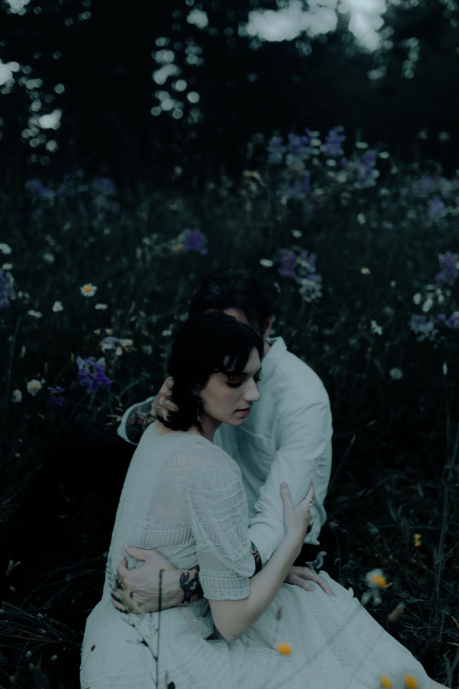 Two people in white clothing sit together among wildflowers at dusk.