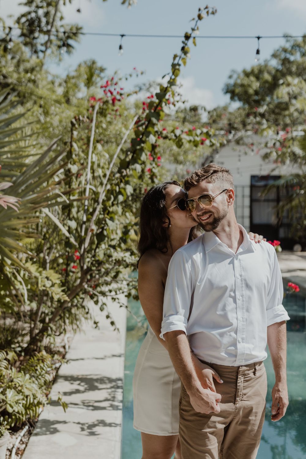 A romantic couple shares an intimate moment among tropical plants and flowers in a sun-drenched courtyard.