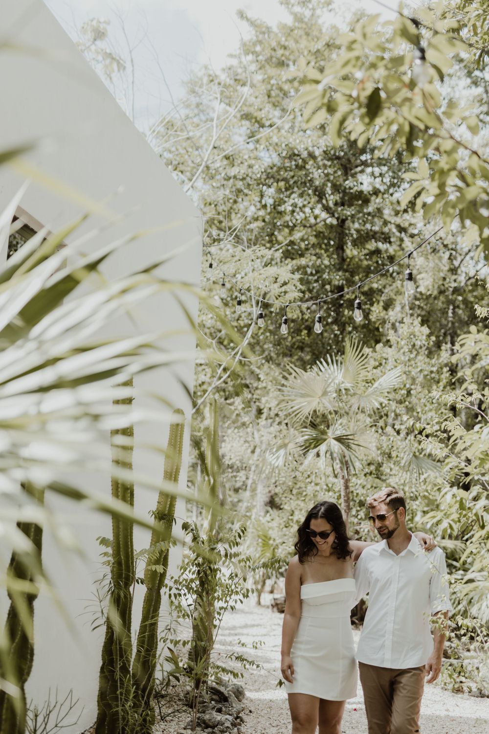 A couple strolls hand in hand along a garden path surrounded by tropical foliage and white walls.