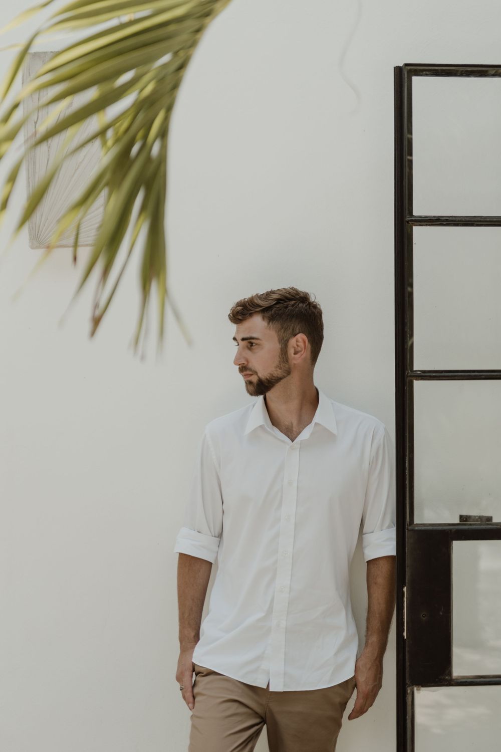 A man in a white shirt poses against a white wall with palm fronds casting shadows.