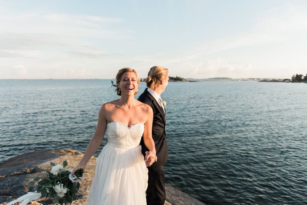 A couple in wedding attire stands on rocky coastal outcrop overlooking calm blue ocean waters at sunset.