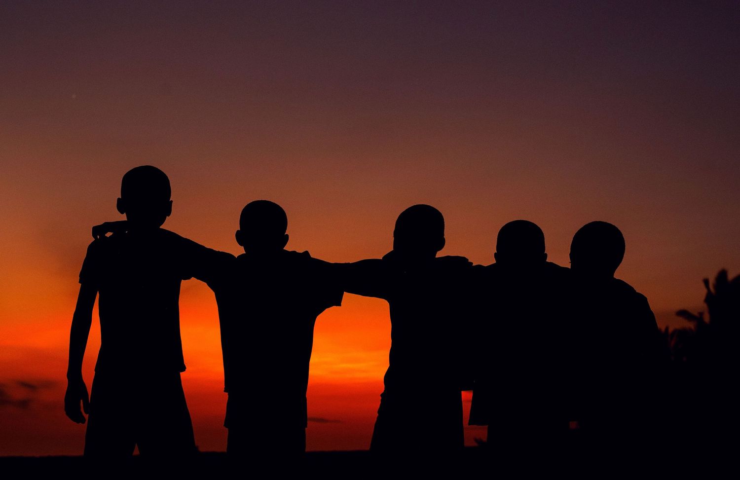 Silhouettes of five people standing together with arms around each other against a vibrant orange sunset sky.