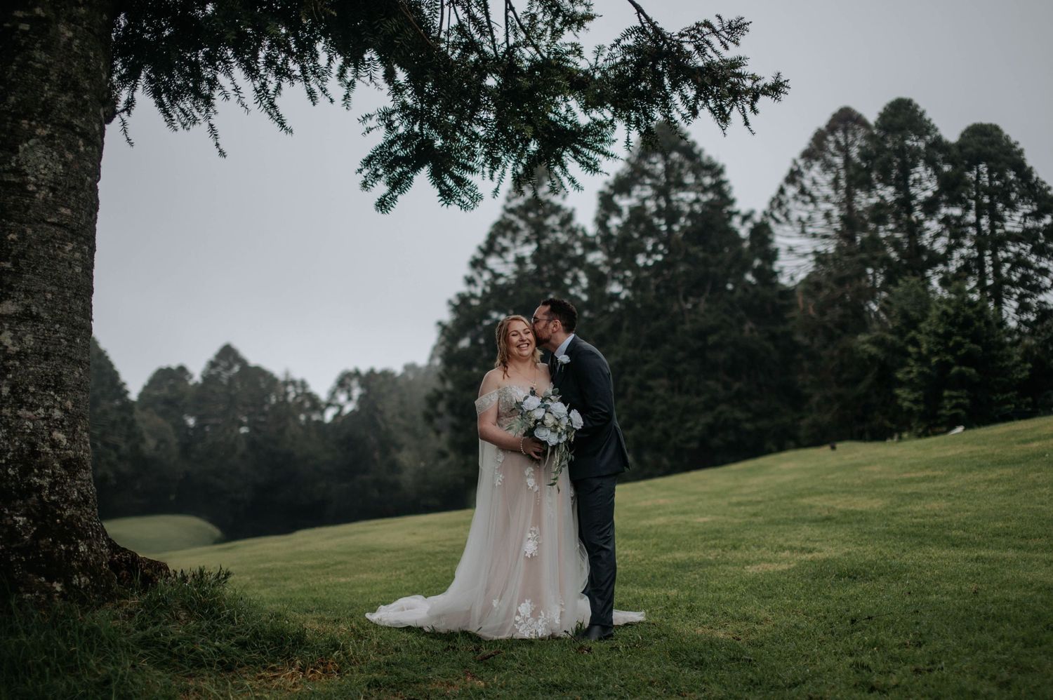 Couple embracing during outdoor wedding photoshoot on misty green hillside.