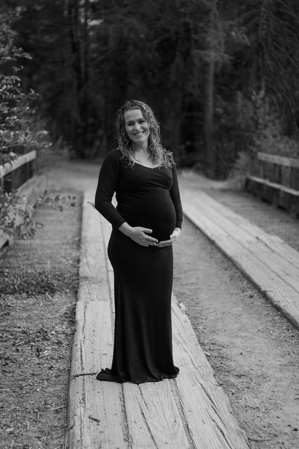 A smiling pregnant woman in a black maternity gown posing on a wooden bridge path.