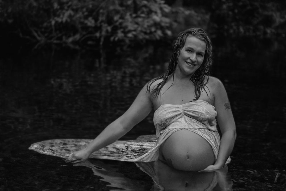 Black and white photo of a pregnant woman sitting in shallow water wearing a white wrap.