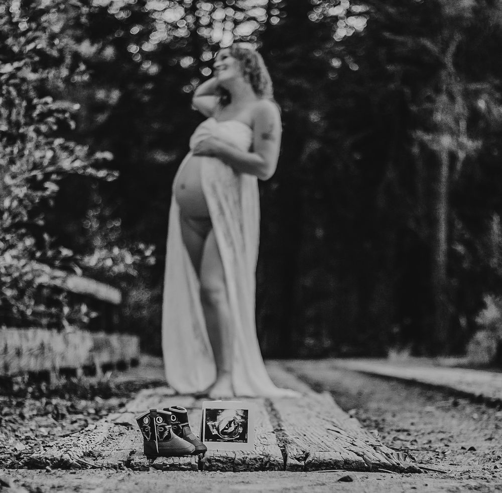 A pregnant woman in a flowing white gown standing on railroad tracks with baby shoes in foreground.