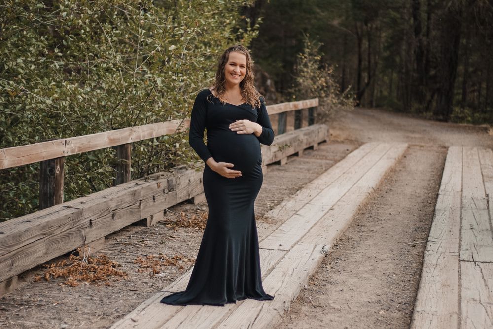 A happy pregnant woman in a dark green maternity gown smiling on a wooden bridge outdoors.