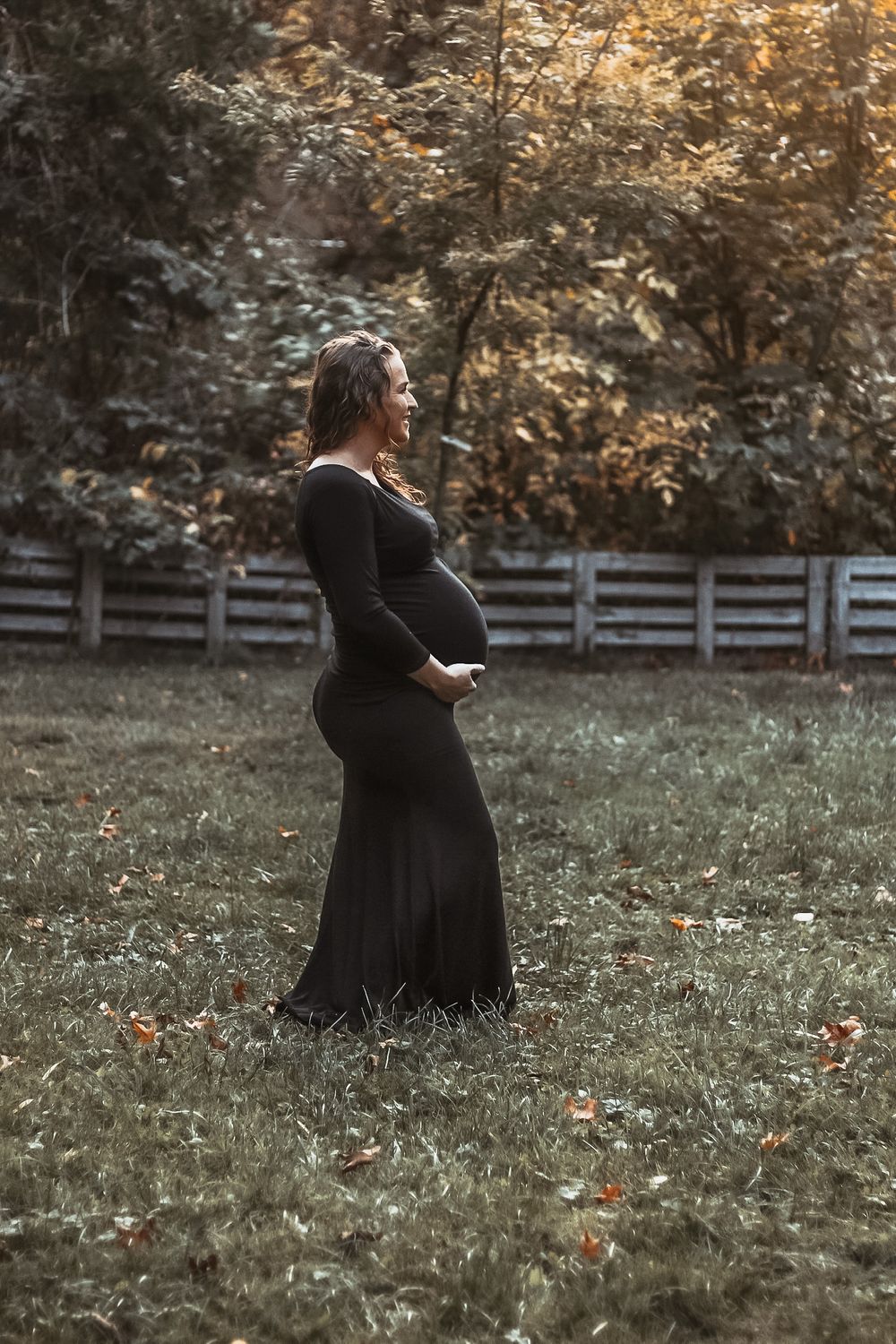 A pregnant woman in a black dress standing in an autumn field beside a white wooden fence.