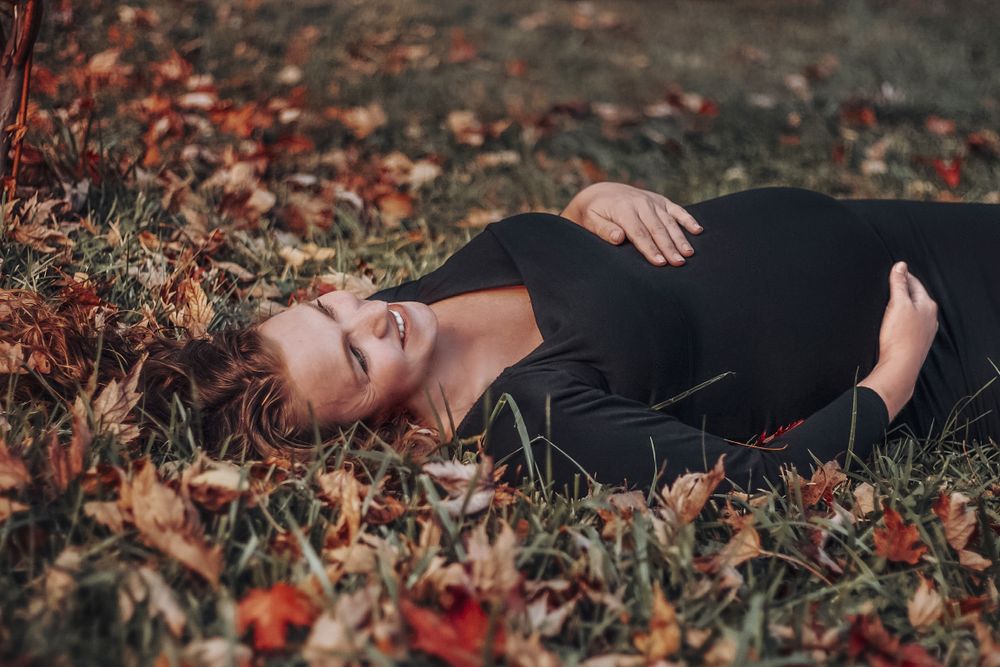 Pregnant woman in black dress lying on autumn leaves in a park during fall season.