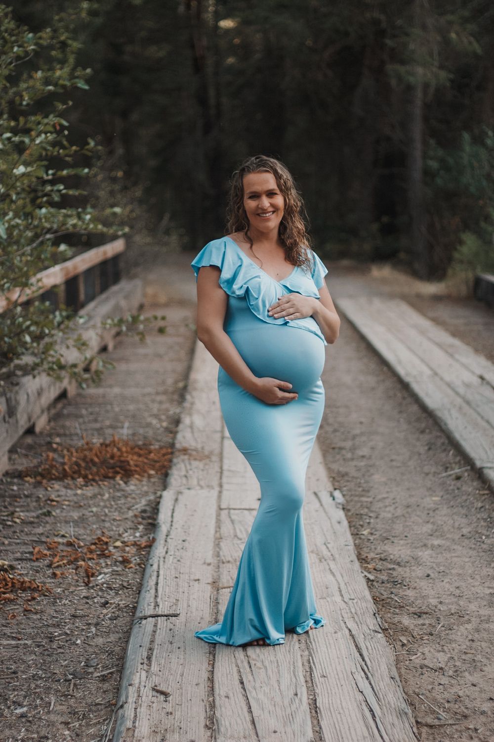 Smiling pregnant woman in light blue maternity gown standing on wooden bridge in forest.